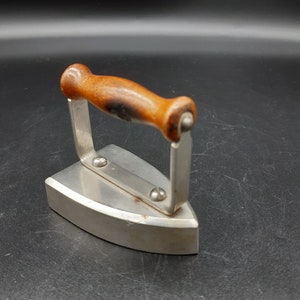 Puede incluir: Una plancha vintage de color plateado con un mango de madera. La plancha tiene una base redondeada y un mango que está unido a la plancha con dos remaches metálicos.