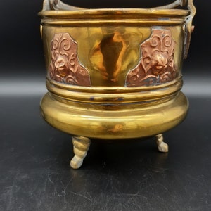 Può includere: Un vaso da fiori vintage in ottone con tre gambe e un design color rame. Il vaso ha una maniglia decorativa e una forma arrotondata.