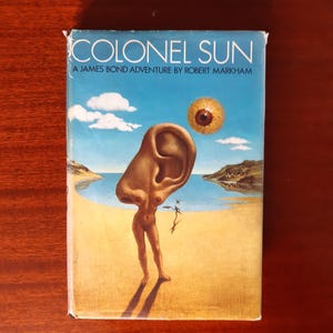 May include: A vintage paperback book cover with the title "Colonel Sun" and the text "A James Bond Adventure by Robert Markham". The cover features a surreal illustration of a woman with a giant ear on her head standing on a beach.
