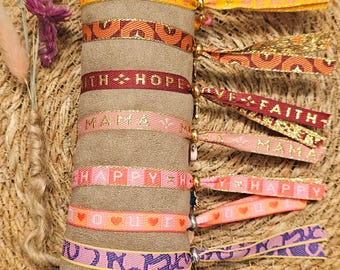 Op de afbeelding: Een set kleurrijke armbanden met gouden letters en verschillende zinnen, waaronder "Love", "Faith", "Hope", "Happy" en "Mama". De armbanden worden gepresenteerd op een bruin suède kussen.