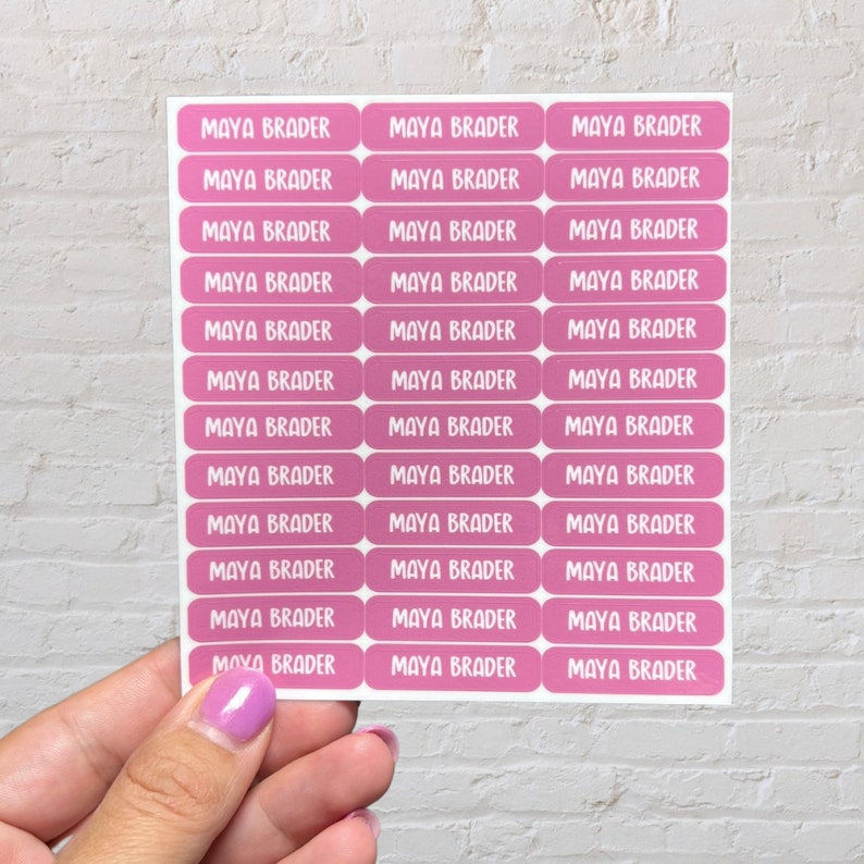 May include: A sheet of pink personalized name labels with the name "MAYA BRADER" printed on each label.