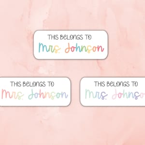 May include: Three white rectangular labels with rounded corners. Each label has the text "THIS BELONGS TO" and the name "Mrs. Johnson" written in rainbow colors.