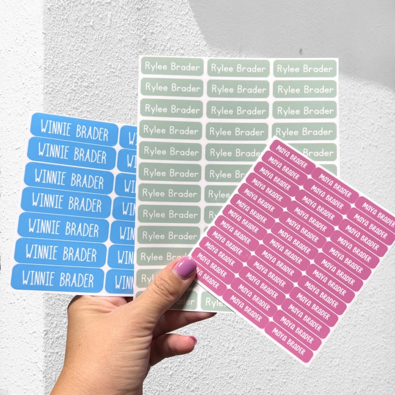 May include: Three sheets of personalized name labels. The first sheet has blue labels with the name "Winnie Brader" printed in white. The second sheet has green labels with the name "Rylee Brader" printed in white. The third sheet has pink labels with the name "Maya Brader" printed in white.