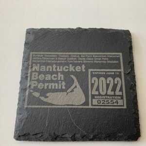 May include: A black slate coaster with a gray inscription that reads "Nantucket Beach Permit" with a map of Nantucket Island and the year "2022" with the text "EXPIRES JUNE 15" and "REGISTRATION 02554".
