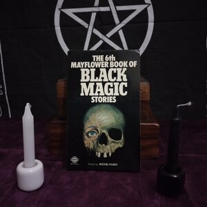 May include: A black book with the title "The 6th Mayflower Book of Black Magic Stories" and a skull and eye illustration on the cover. The book is sitting on a wooden surface with two candles, one white and one black, on either side.