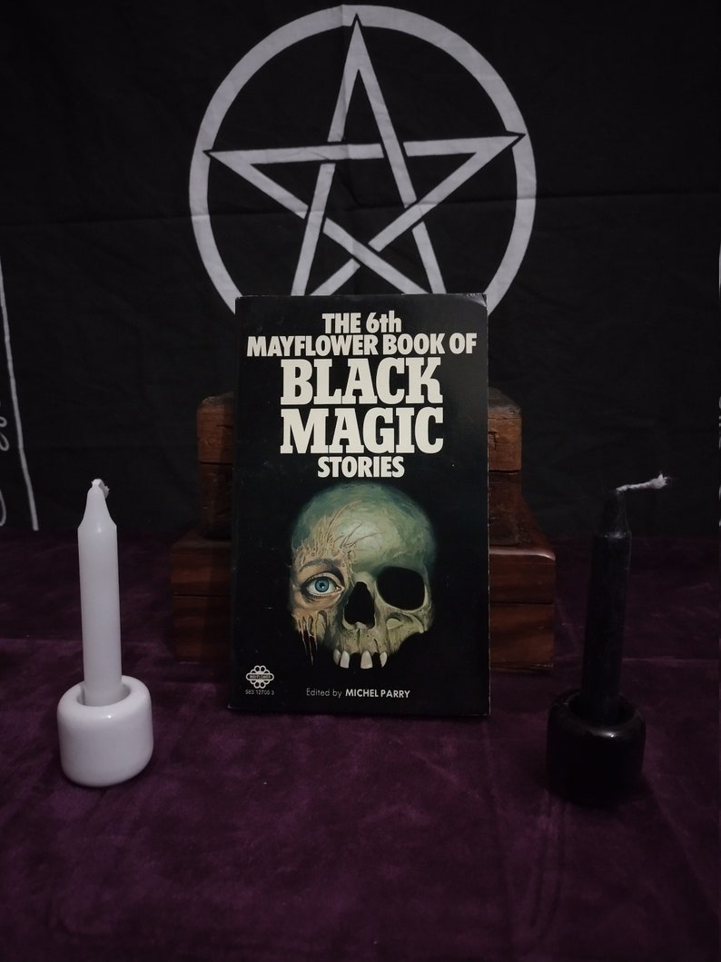 May include: A black book with the title "The 6th Mayflower Book of Black Magic Stories" and a skull and eye illustration on the cover. The book is sitting on a wooden surface with two candles, one white and one black, on either side.
