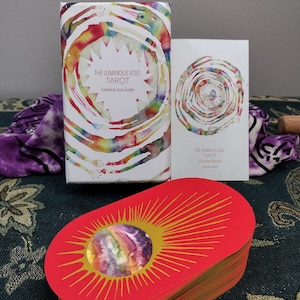 May include: A tarot deck with a red card design featuring a sunburst pattern and a central watercolor image. The deck includes a box and guide book, both with watercolor designs and the text "THE LUMINOUS VOID TAROT".