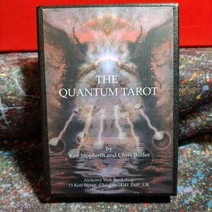 May include: A black tarot card box with the title "The Quantum Tarot" in white text. The box features an abstract image with a central light source and connecting orbs. The authors' names and the bookshop address are also visible.