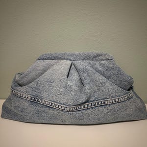 May include: A light blue acid-wash denim clutch bag featuring a gathered, pleated top design with a structured frame closure and a horizontal seam detail across the front body of the pouch.