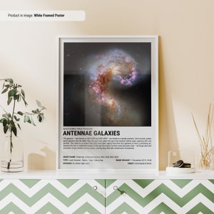 May include: White framed poster featuring the Antennae Galaxies, a pair of interacting galaxies. The image shows swirling formations of stars and gas, with text at the bottom identifying the galaxies and their characteristics. The poster is displayed on a white cabinet.