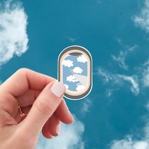 May include: A sticker featuring an airplane window design with clouds and blue sky. The sticker is held against a backdrop of a bright blue sky filled with fluffy white clouds. The sticker has a white border.