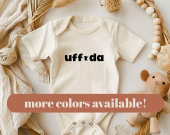 May include: A cream-colored short-sleeve baby bodysuit featuring the black text "uff da" with the letter "i" replaced by a small map outline of the state of Minnesota. The garment has a round neckline and three metal snaps at the bottom.