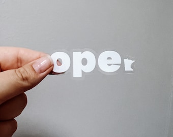 Op de afbeelding: Een hand houdt een witte vinyl sticker vast met de tekst "open" op een grijze achtergrond.