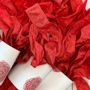 May include: A close-up of a pile of red silk ribbon, wrapped around two white labels with a pink rose design. The ribbon is soft and delicate, perfect for crafting and decorating.