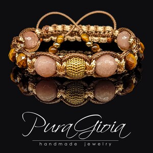 May include: A brown and beige braided bracelet with gold and brown beads. The bracelet has a sunstone and tiger's eye beads. The bracelet is on a black background. Pura Gioia handmade jewelry.