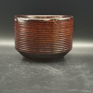 May include: A cylindrical ceramic bowl with a dark brown, glossy glaze and a series of horizontal, raised ridges encircling the exterior. The bowl features a wide, open top and a slightly tapered base, showcasing a uniform, textured surface throughout.