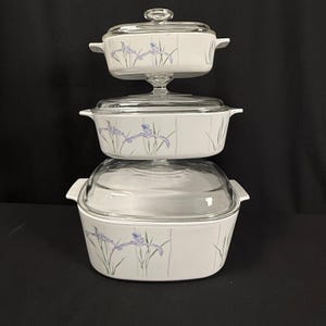 May include: Three-piece white casserole dish set with glass lids. Each dish features a floral iris design in purple and green. The dishes are stacked, with the smallest on top and the largest on the bottom. The set is on a black background.