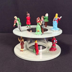 May include: A vintage carousel figurine with miniature figures of people in various outfits. The figures are arranged on a white circular base with a clear plastic cylinder in the center. The figures include a violinist, dancers, and people carrying luggage.