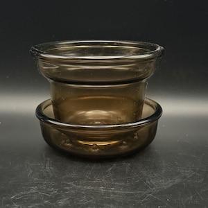 May include: A translucent dark brown glass planter pot with a matching circular saucer base. The planter features a wide, rounded rim and a tapered body, while the saucer has a shallow, curved design with small raised bumps on the bottom surface.