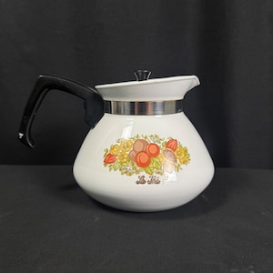 May include: A white teapot with a black handle and lid, featuring a metallic band around the neck. The body displays a colorful botanical illustration of fruits and leaves with the text Le Thé printed in gold script below the design.