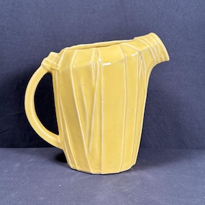 May include: A yellow ceramic pitcher with a curved handle and a spout. The pitcher has a ribbed design and a glossy finish. The pitcher is a light yellow color and is likely used for serving beverages. The pitcher is approximately 8 inches tall.