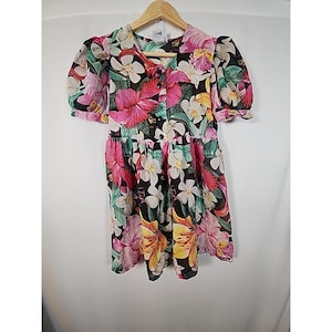 May include: A vintage floral print dress with a black background, featuring large pink, white, and yellow flowers. The dress has short, puffed sleeves and a button-down front. The dress is hanging on a wooden hanger.