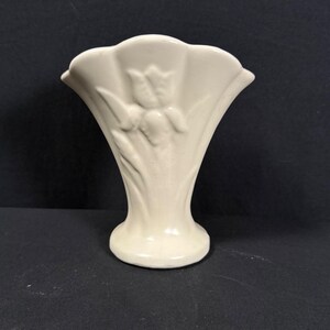 May include: A cream-colored ceramic vase with a flared, scalloped rim and a raised, embossed floral design featuring a central tulip motif. The vase has a smooth, glossy finish and a circular base, standing upright with a symmetrical silhouette.