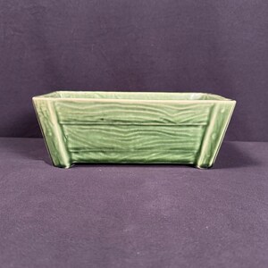 May include: A rectangular, light green ceramic planter with a wood-grain texture. The planter has a slightly trapezoidal shape, with angled sides and a flat base. The overall design is simple and rustic, suitable for small plants or succulents.