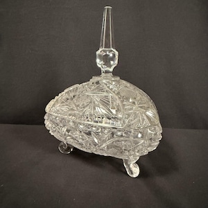 May include: Clear, cut-glass candy dish with a pointed finial on the lid. The dish has a rounded, oval shape and sits on three small, curved feet. The glass is intricately patterned with geometric and floral designs.