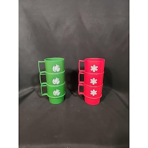 May include: Two stacks of plastic mugs. One stack is green with a white floral design, and the other is red with a white snowflake design. Each mug has a handle and is stacked on top of each other.