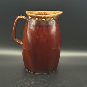 May include: A ceramic pitcher with a glossy, deep brown glaze and a lighter, mottled tan drip pattern around the rim. The vessel features a curved handle on the left side and a wide, rounded body that tapers slightly toward the top opening.