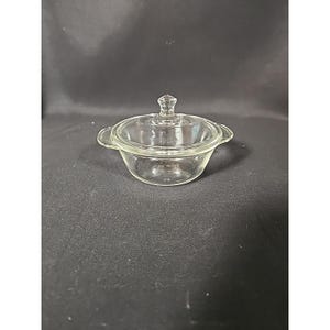 May include: A clear glass casserole dish with a lid. The dish has two small handles and a knob on the lid. The dish is sitting on a black surface.