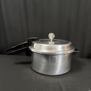 May include: A silver-colored aluminum pressure cooker with a large black plastic handle on the left side and a smaller black handle on the right. The lid features a central circular metal pressure regulator valve and a locking mechanism.