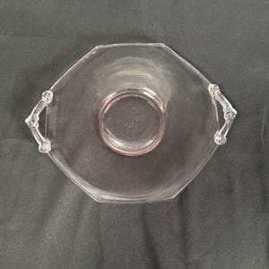 May include: A vintage pink glass serving dish with an octagonal shape and two decorative handles. The dish has a central circular indentation and is made of transparent glass.
