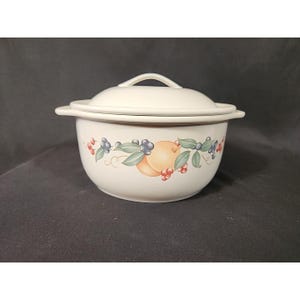 May include: A white ceramic bowl with a matching lid. The bowl features a painted design of peaches, blueberries, and red berries with green leaves. The lid has a small handle for easy removal. The bowl is likely for serving or storage.
