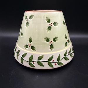 May include: A tapered ceramic lamp shade featuring a pale green upper section decorated with embossed green holly leaves and red berries. The lower border is white and displays a continuous vine pattern with green leaves and small red berries.