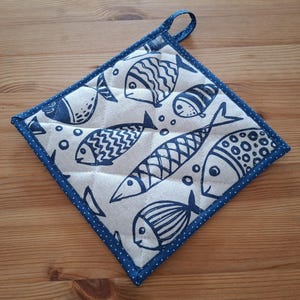 May include: Square quilted pot holder featuring a blue fish design on a cream backdrop. It has a blue polka dot border and a hanging loop. The fish illustrations are diverse in style and size, adding visual interest.