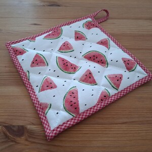 Può includere: Un sottopentola quadrato e trapuntato con sfondo bianco con fette di anguria e semi neri. I bordi sono rifiniti con un motivo a quadretti rosso e bianco. Un piccolo anello è attaccato per appenderlo.