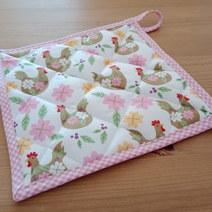 Könnte beinhalten: Ein rosa-weiß gesteppter Topflappen mit einem karierten Rand. Der Topflappen zeigt ein sich wiederholendes Muster aus braunen Hühnern und rosa Blumen.