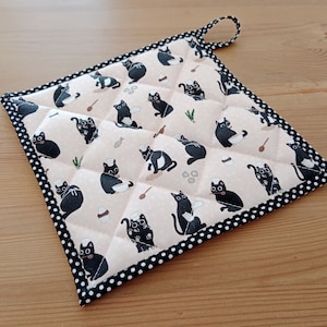 Könnte beinhalten: Ein schwarzer und weißer gesteppter Topflappen mit schwarzem und weißem Rand. Der Topflappen ist mit einem Muster aus schwarzen Katzen verziert, die Kochmützen tragen.