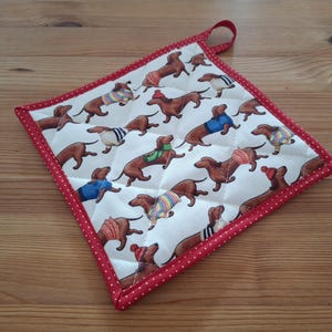 Könnte beinhalten: Ein quadratischer, gesteppter Topflappen mit einem sich wiederholenden Muster von Dackeln in Pullovern und Hüten. Die Hunde sind braun mit bunten Outfits. Der Topflappen hat einen roten Pünktchenrand und eine rote Schlaufe.