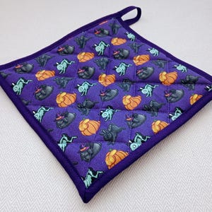 Könnte beinhalten: Quadratischer, gesteppter Topflappen in Lila mit einer Aufhängeschlaufe. Der Stoff ist mit orangefarbenen Kürbissen, schwarzen Hexenkatzen und türkisfarbenen Skelettfiguren gemustert. Dunkelviolette Kanten.