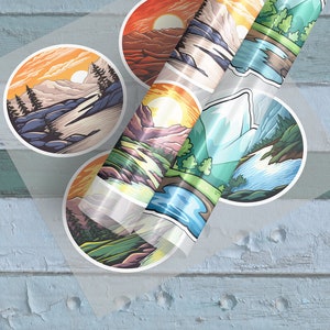 May include: A sheet of colorful mountain landscape stickers. The stickers are round and feature different scenes of mountains, rivers, and sunsets. The stickers are on a clear sheet and are ready to be peeled off and used.