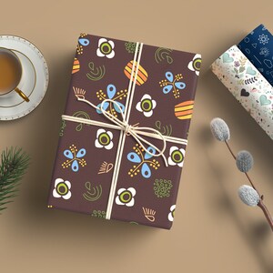 May include: A brown gift box wrapped in paper with a colorful floral pattern. The paper features white, blue, yellow, orange, and green flowers and abstract shapes. The box is tied with a white string.