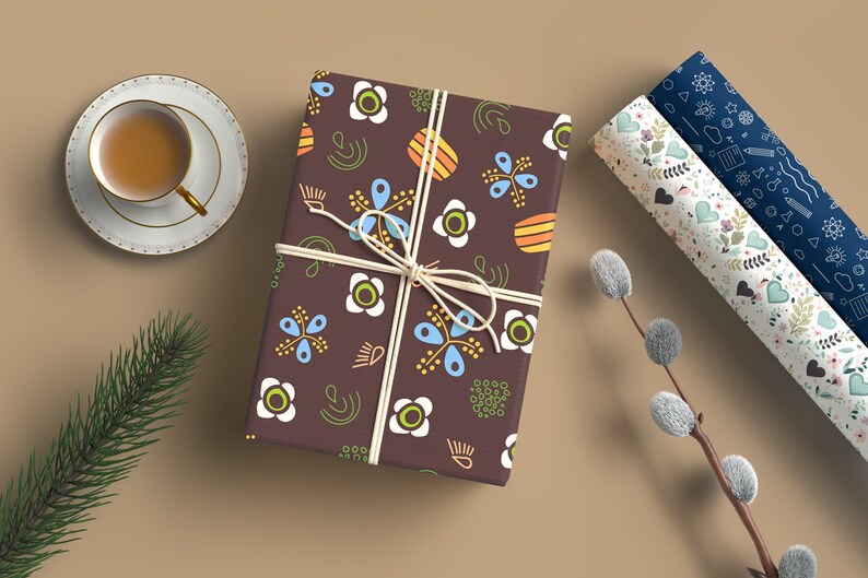 May include: A brown gift box wrapped in paper with a colorful floral pattern. The paper features white, blue, yellow, orange, and green flowers and abstract shapes. The box is tied with a white string.