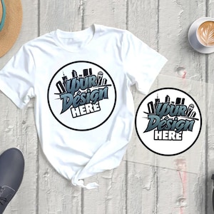 Peut inclure: T-shirt blanc avec un motif circulaire indiquant "Your Design HERE" dans une police stylisée. Le motif présente une silhouette de paysage urbain. Le t-shirt est à côté d'une version plus petite du motif, d'un chapeau, de chaussures et d'une tasse de café.