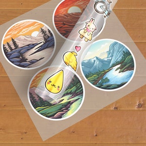 May include: A sheet of clear sticker paper with five stickers. The stickers are round and feature colourful illustrations of landscapes and a cartoon character. One sticker shows a mountain range with a river flowing through it. Another sticker shows a sunset over a mountain range. A third sticker shows a cartoon character with a heart above its head. The fourth sticker shows a cartoon character with a yellow drop of liquid above its head. The fifth sticker shows a cartoon character with a yellow drop of liquid above its head.