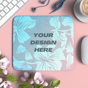 Puede incluir: Una alfombrilla de ratón con estampado floral azul y blanco con el texto "YOUR DESIGN HERE" sobre una superficie rosa con un teclado blanco, un ratón blanco, una taza de café y unos auriculares.