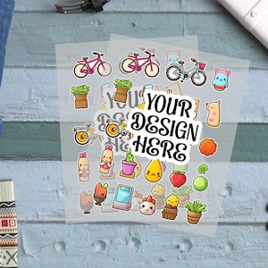 Puede incluir: Una hoja de papel adhesivo transparente con pegatinas de dibujos animados de colores. Las pegatinas incluyen bicicletas, plantas, frutas y otros diseños bonitos. El texto "YOUR DESIGN HERE" está impreso en la hoja de pegatinas.