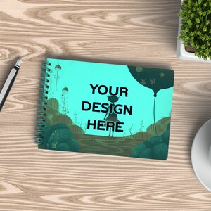 Puede incluir: Una libreta de espiral azul turquesa con una ilustración caprichosa de una niña de pie en un campo con un globo aerostático. El texto "YOUR DESIGN HERE" está impreso en negro en la portada.